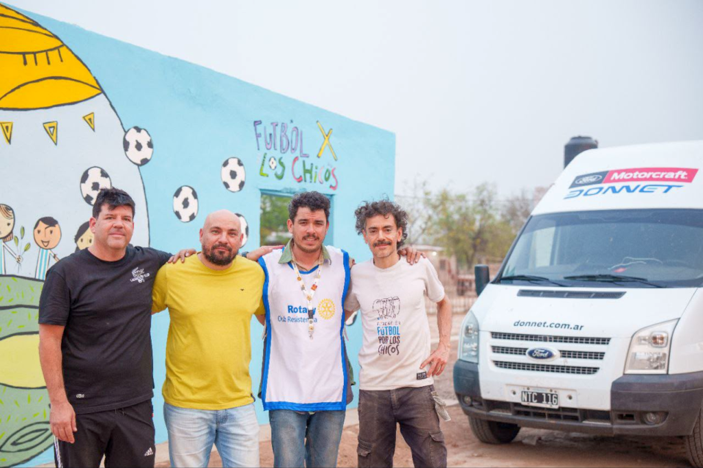 Cuatro voluntarios frente al mural celeste "Fútbol por los Chicos" del albergue de Tres Pozos, uno con chaleco de Rotary Club Resistencia, junto a la camioneta del operativo