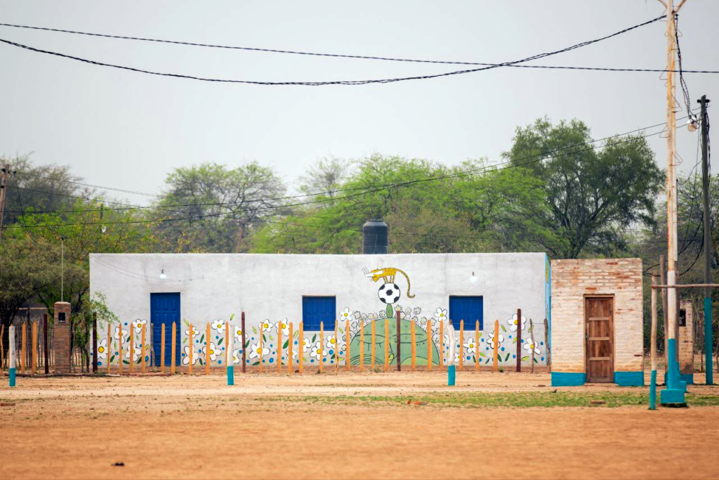 Frente del albergue estudiantil de Tres Pozos: edificio blanco con puertas y ventanas azules, mural de un yaguareté con pelota de fútbol y cerco de palos pintados, en el Impenetrable chaqueño