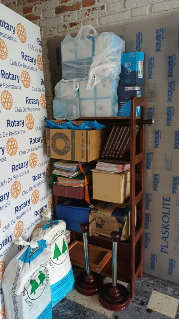 Estantería con las donaciones ordenadas para la escuela de Isla Soto: kits escolares, libros, galletitas y materiales, junto al banner de Rotary Club Resistencia