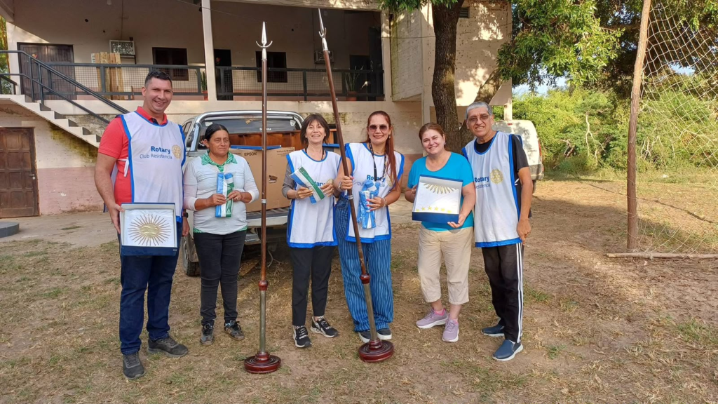 Socios de Rotary Club Resistencia junto a una integrante de la comunidad de Isla Soto con las banderas de ceremonia y los mástiles donados, frente a la Escuela N.º 797